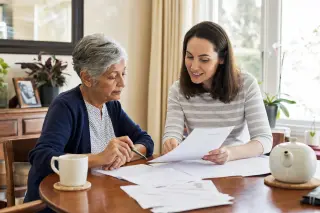 Senior person and supportive family member filling out paperwork together at a sunlit kitchen table in a cozy apartment.